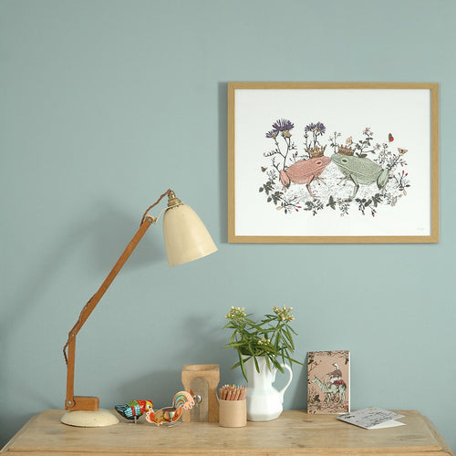 Framed print of two kissing frogs illustration on wall behind desk